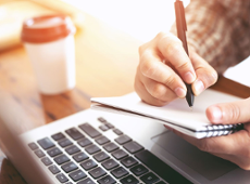 na stole leży otwarty komputer i kubek z kawą na wynos a na pierwszym planie ręce notujące coś w notesie