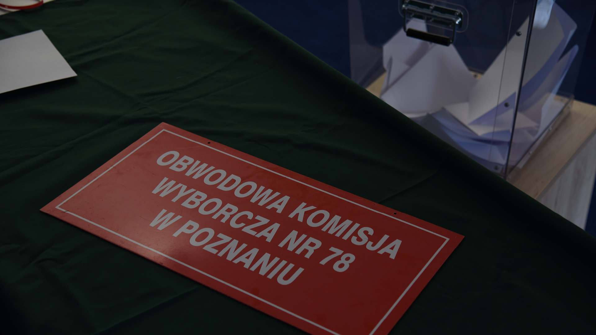 na stole nakrytym zielonym suknem leży czerwona prostokątna tablica informacyjna z białym napisem obwodowa komisja wyborcza nr 78 w Poznaniu. W tle widać wypełnioną do połowy kartami wyborczymi przeźroczystą urnę wyborczą.
