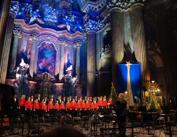 Na stopniach ołtarza stoi liczny chór złożony z chłopców i młodych mężczyzn. Młodsi chórzyści ubrani są w charakterystyczne czerwone swetry, natomiast starsi występują w eleganckich czarnych garniturach z muszkami. Przed nimi stoi dyrygent, widoczny od tyłu, kierujący występem.