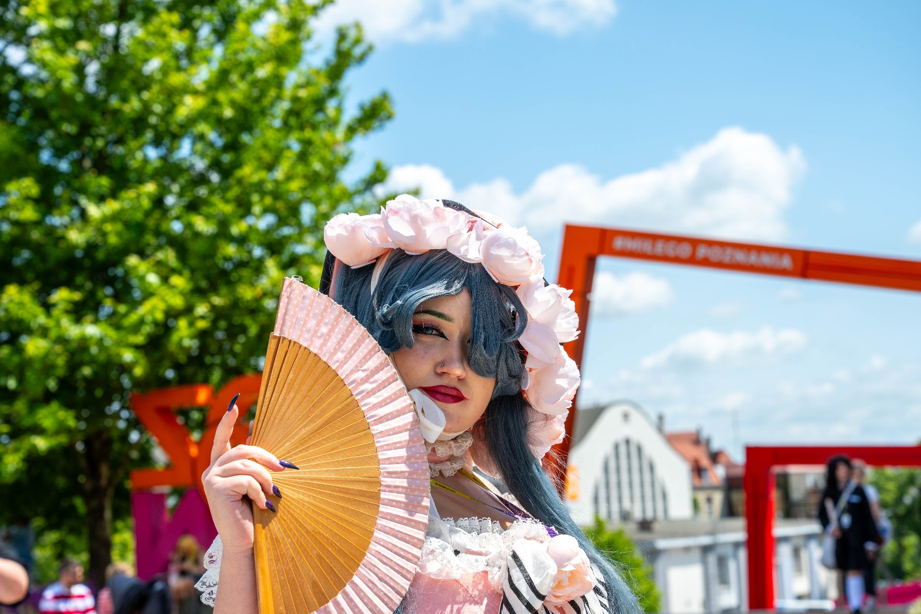 Na zdjęciu cosplayerka w różowej peruce, za nią bramka z napisem: miłego Poznania