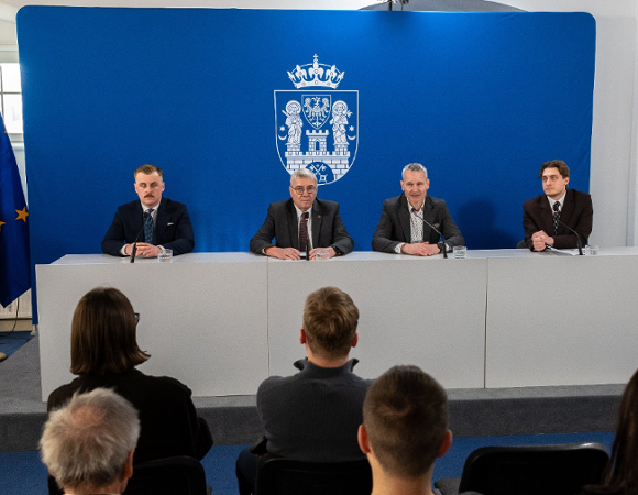 Na zdjęciu czterej mężczyźni siedzący za stołem konferencyjnym, w tle ścianka z herbem miasta