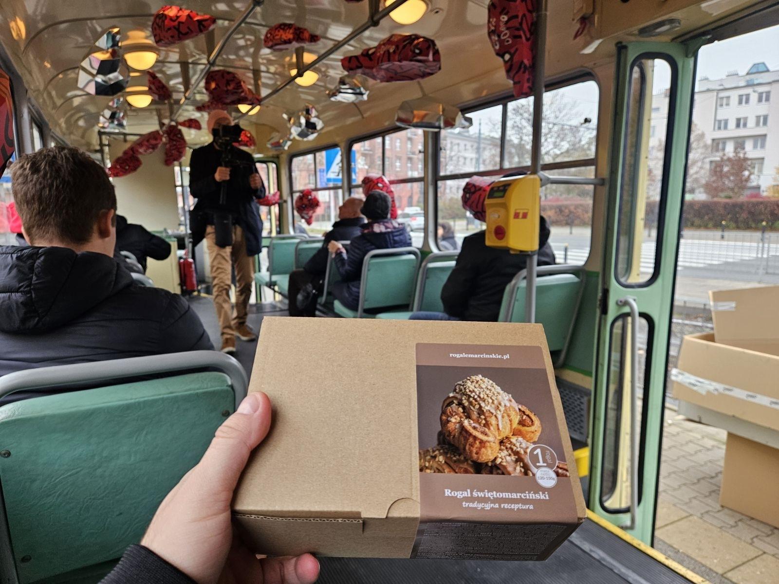 Na zdjęciu dłoń trzymająca rogal, w tle domki budnicze