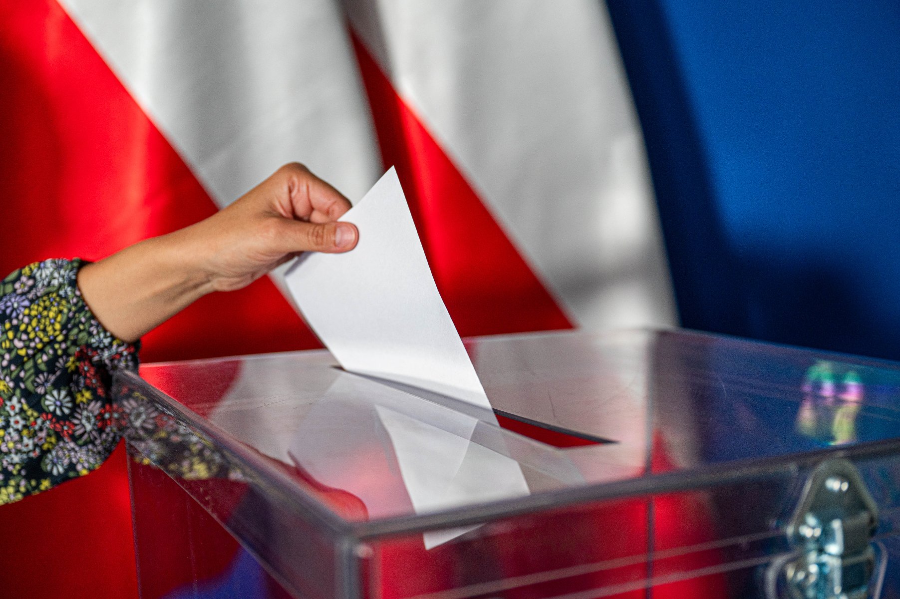 Na zdjęciu dłoń wrzucająca głos do urny, w tle dwie flagi Polski