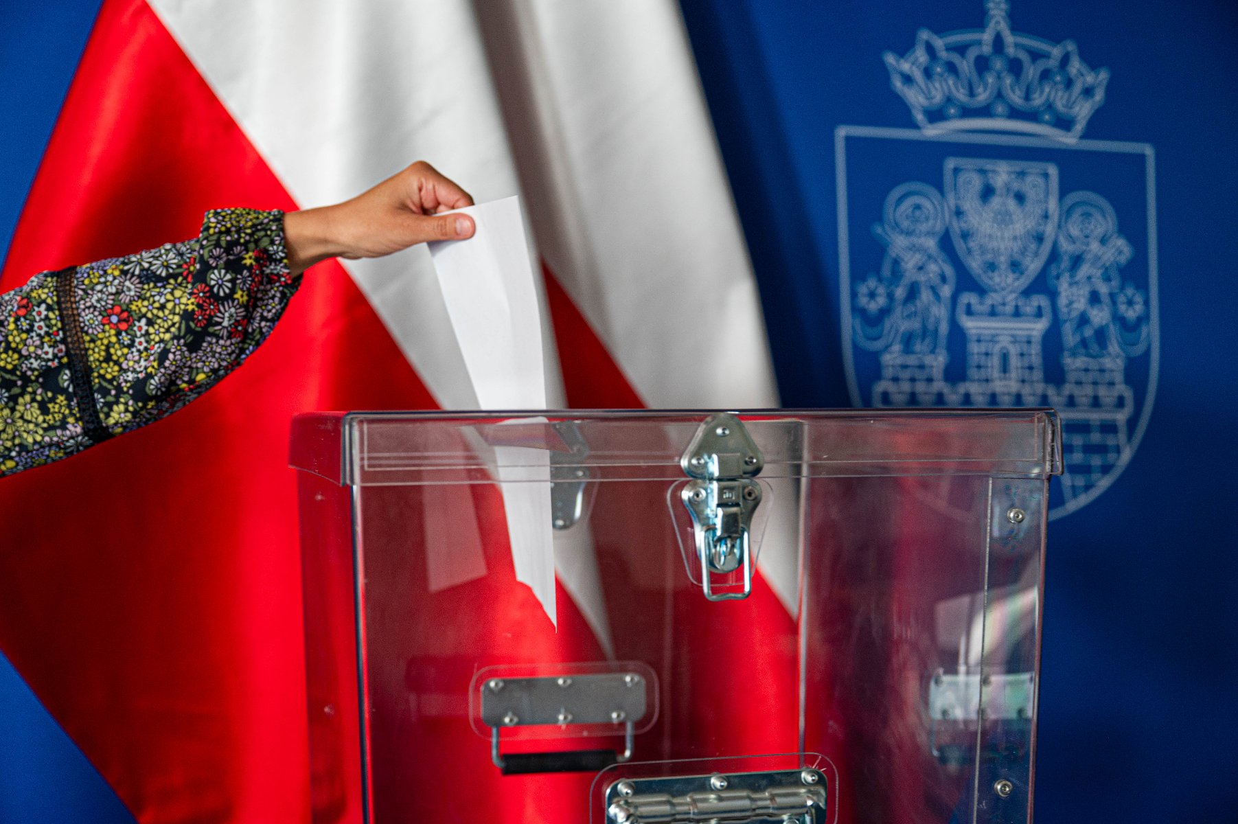 Na zdjęciu dłoń wrzucająca głos do urny, w tle dwie flagi Polski i herb Miasta