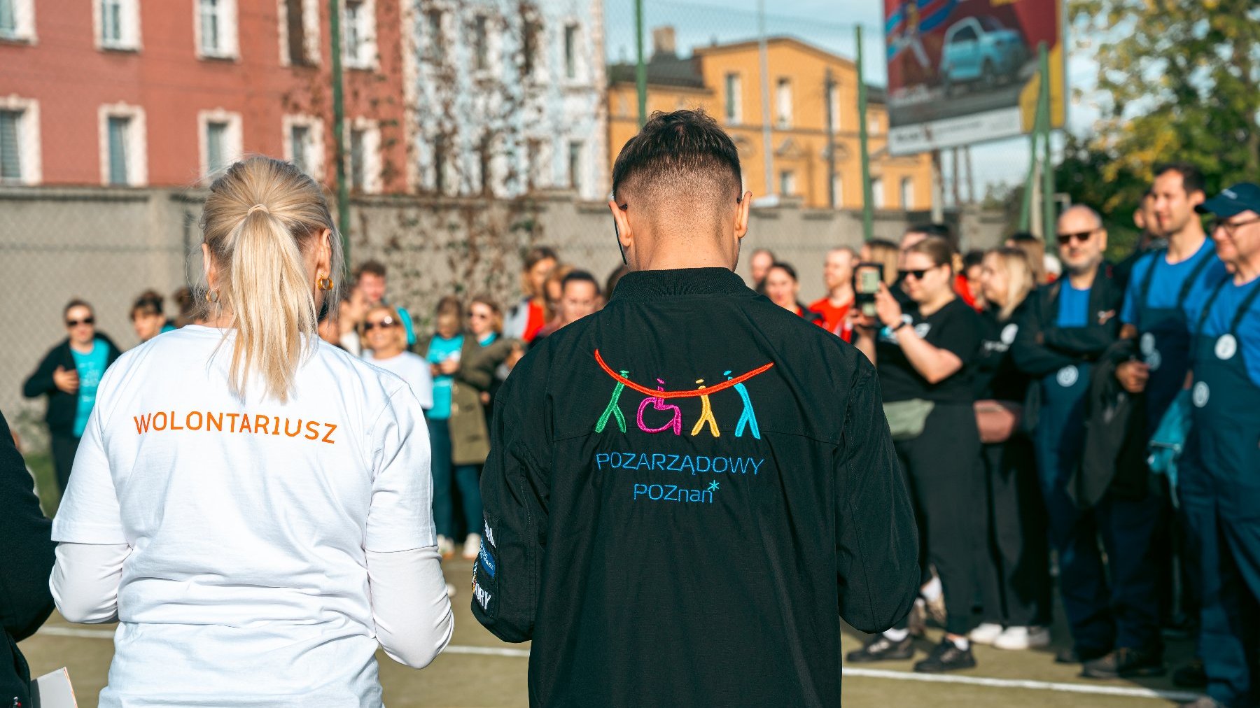 Na zdjęciu dużo ludzi, dwie osoby stoją tyłem do obiektywu, mają koszulki z napisem wolontariusz