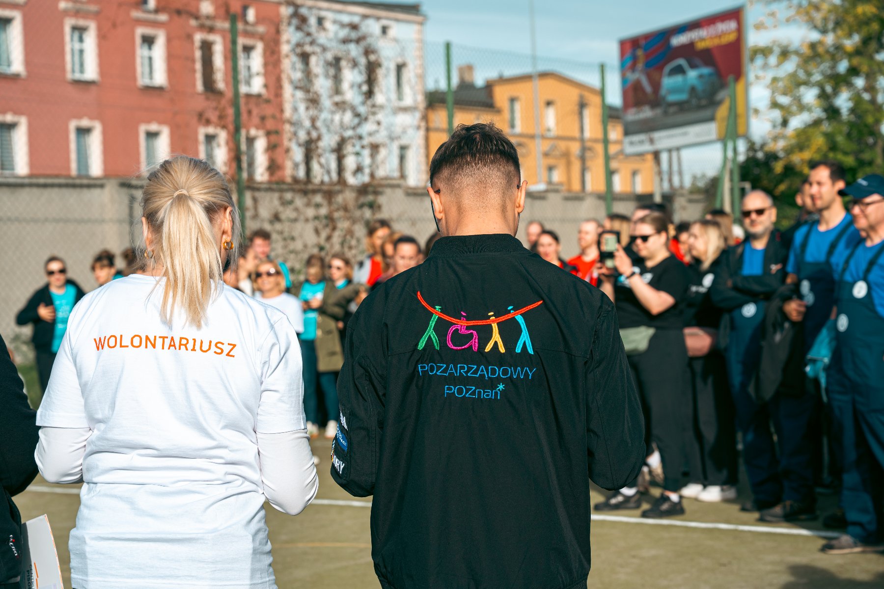 Na zdjęciu dużo ludzi, dwie osoby stoją tyłem do obiektywu, mają koszulki z napisem wolontariusz