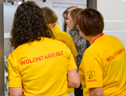 Na zdjeciu dwie kobiety stojące tyłem, na żółtych koszulkach mają napis: wolontariusz