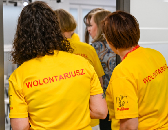 Na zdjeciu dwie kobiety stojące tyłem, na żółtych koszulkach mają napis: wolontariusz