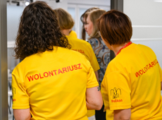 Na zdjeciu dwie kobiety stojące tyłem, na żółtych koszulkach mają napis: wolontariusz