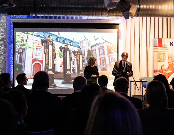 Na zdjęciu dwie osoby na scenie, przed nimi publicznosć, za nimi ekran