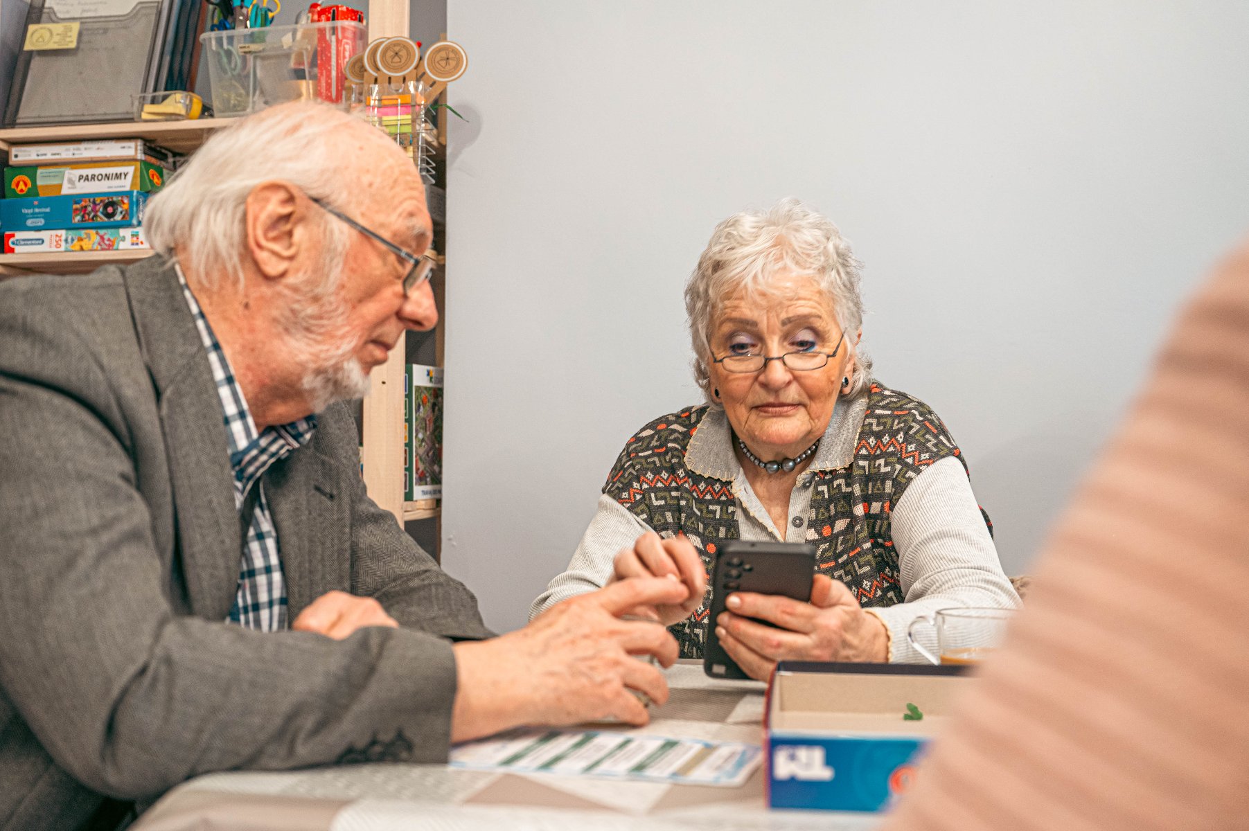 Na zdjeciu dwoje seniorów przy stoliku, jedna osoba trzyma w ręku smartfon