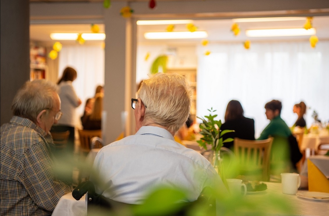 Na zdjęciu jasna sala, na pierwszym planie dwóch seniorów tyłem do obiektywu, w tle inne osoby