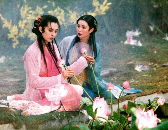 Na zdjęciu kard z filmu, dwie kobiety w tradycyjnych strojach Azji rozmawiają na polu pełnym kwiatów