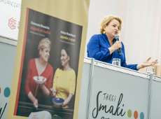 Na zdjęćiu Katarzyna Bosacka mówiąca ze sceny do mikrofonu. Z boku stoi rollup