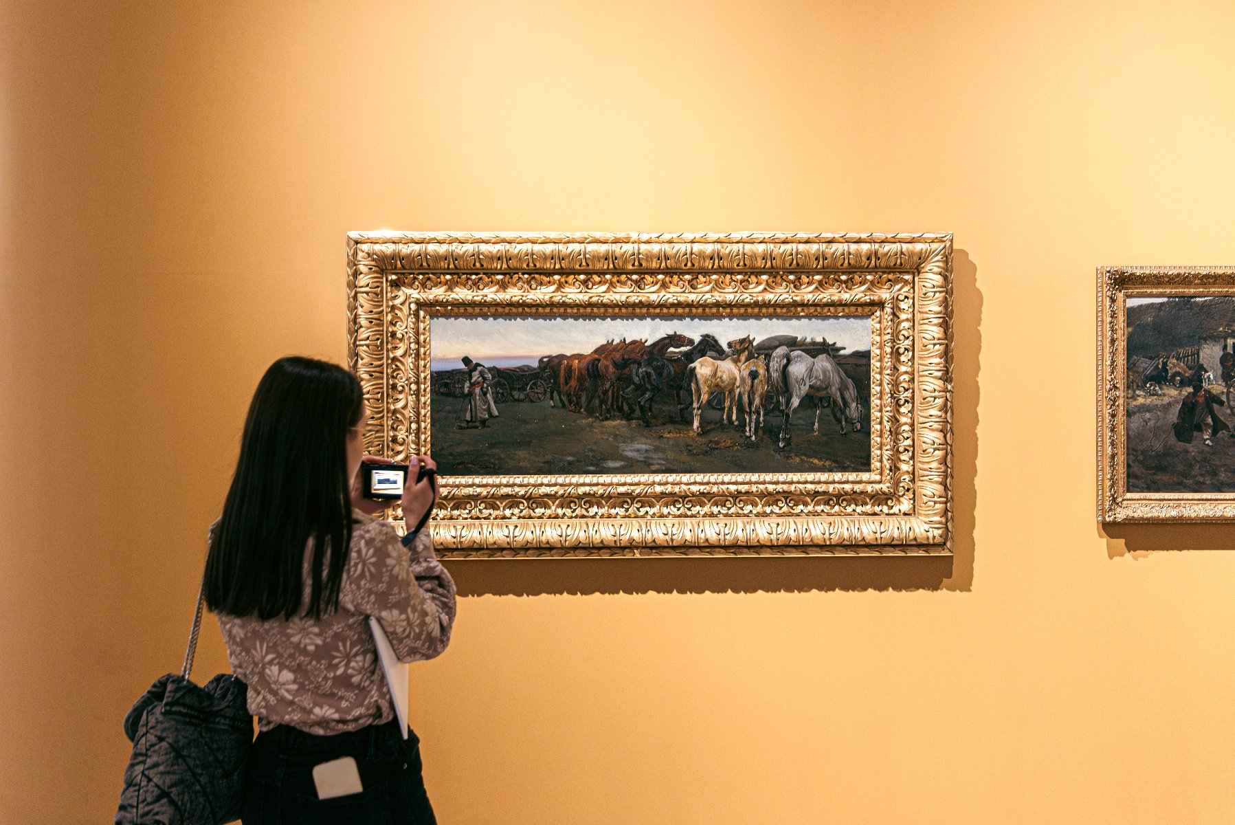 Na zdjęciu kobieta robiąca zdjęcie obrazowi wiszącemu na żółtej ścianie