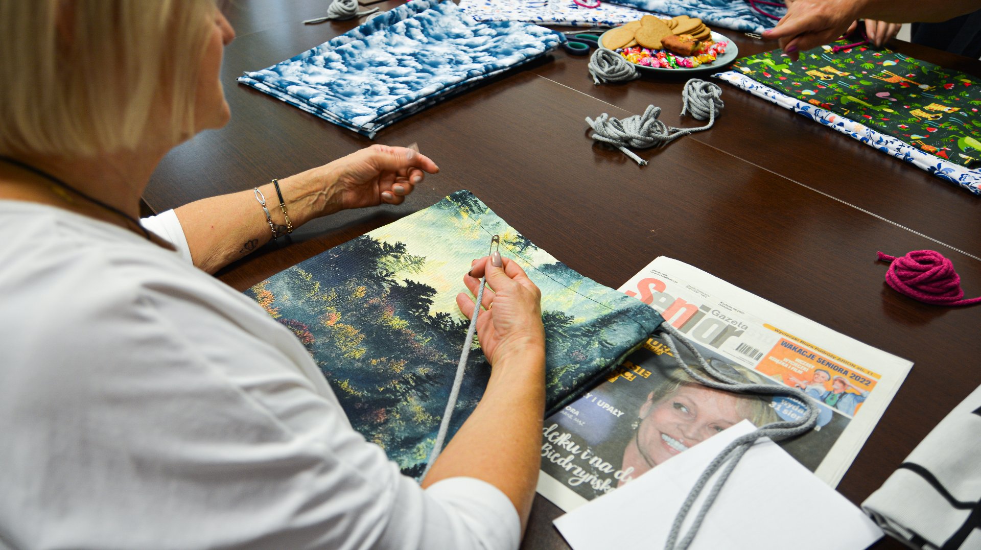 Na zdjęciu kobieta siedząca przy stole, przed nią tkanina i gazeta z napisem senior