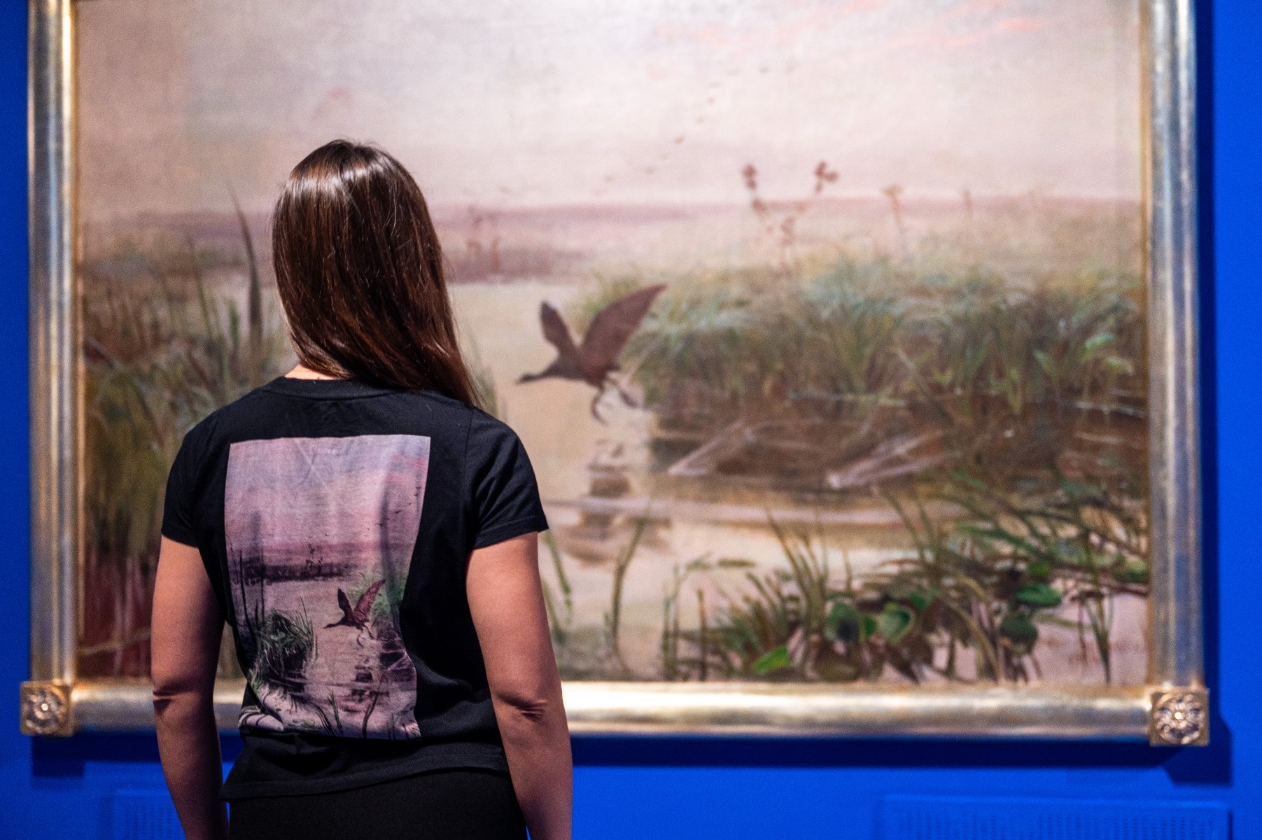 Na zdjęciu kobieta stojąca przed obrazem Chełmońskiego, nosi koszulkę, na której nadrukowany jest ten sam obraz