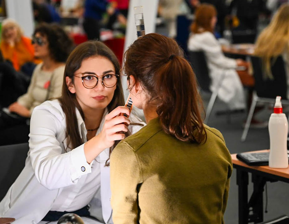 Na zdjęciu kobieta w medycznym fartuchu przytyka coś do twarzy kobiecie siedzącej tyłem do obiektywu