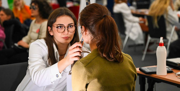 Na zdjęciu kobieta w medycznym fartuchu przytyka coś do twarzy kobiecie siedzącej tyłem do obiektywu