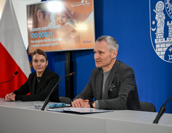 Na zdjęciu mężczyzna i kobieta za stołem konferencyjnym, za nimi ścianka z herbem miasta oraz grafika przedstawiająca dziecko