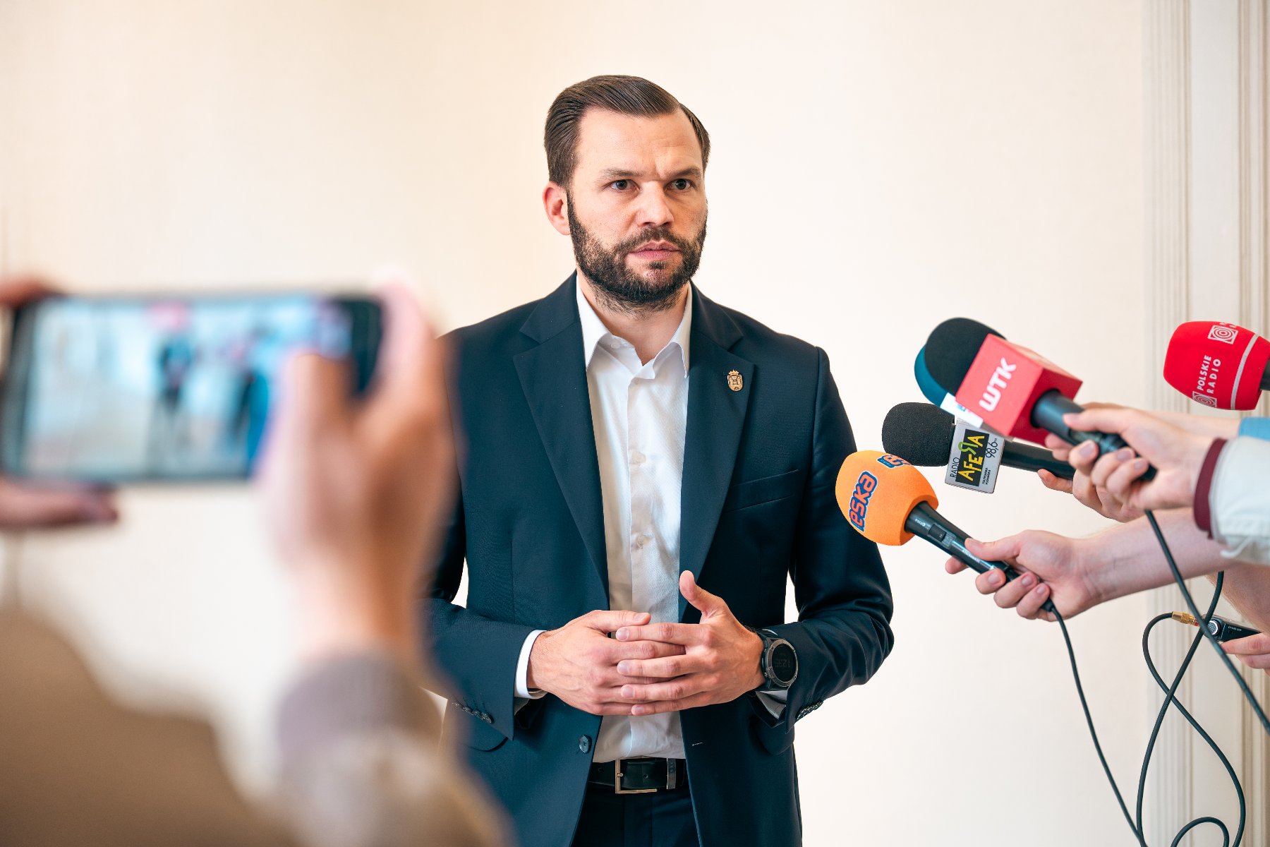 Na zdjęciu mężczyzna w garniturze, mówi coś do mikrofonów, które trzymają niewidoczni w kadrze dziennikarze