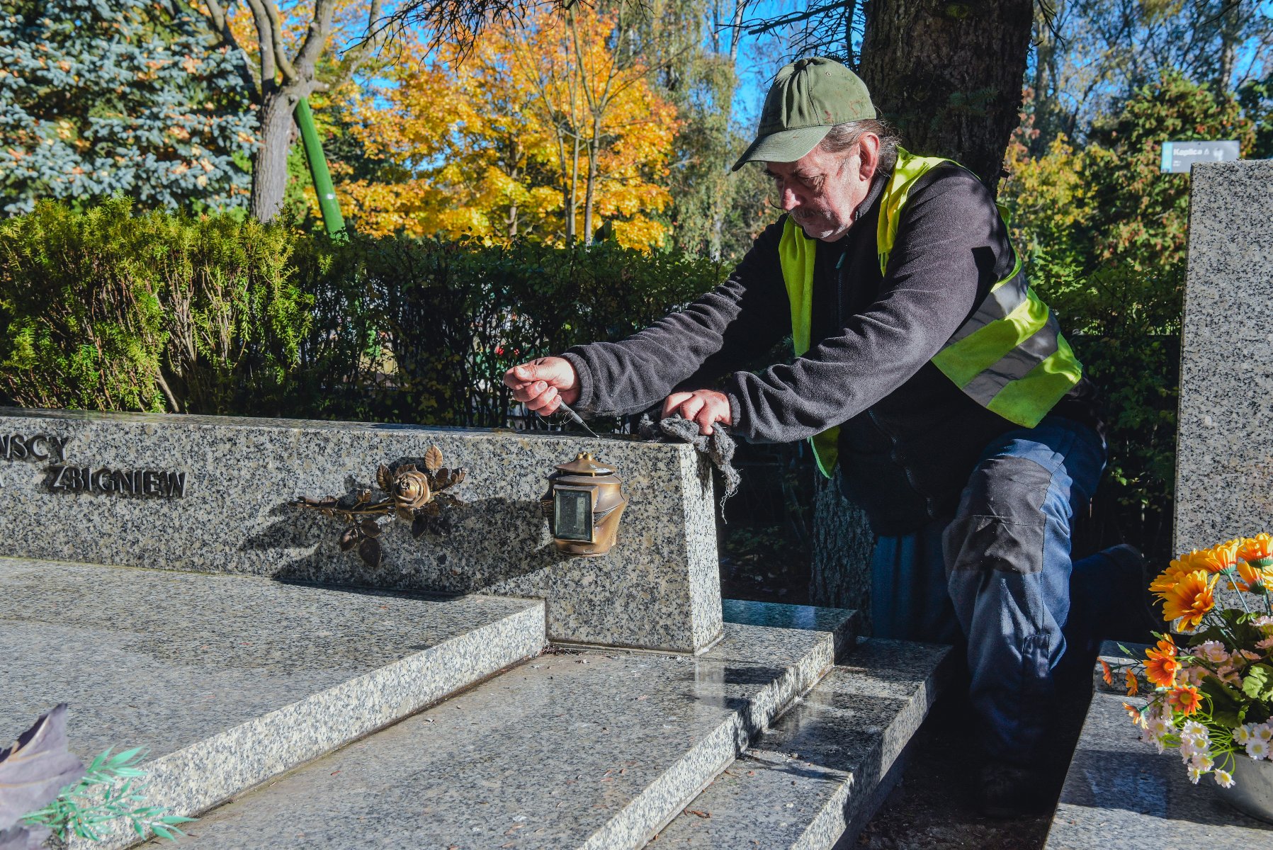 Na zdjęciu mężczyzna w odblaskowej kamizelce, sprzątający nagrobek