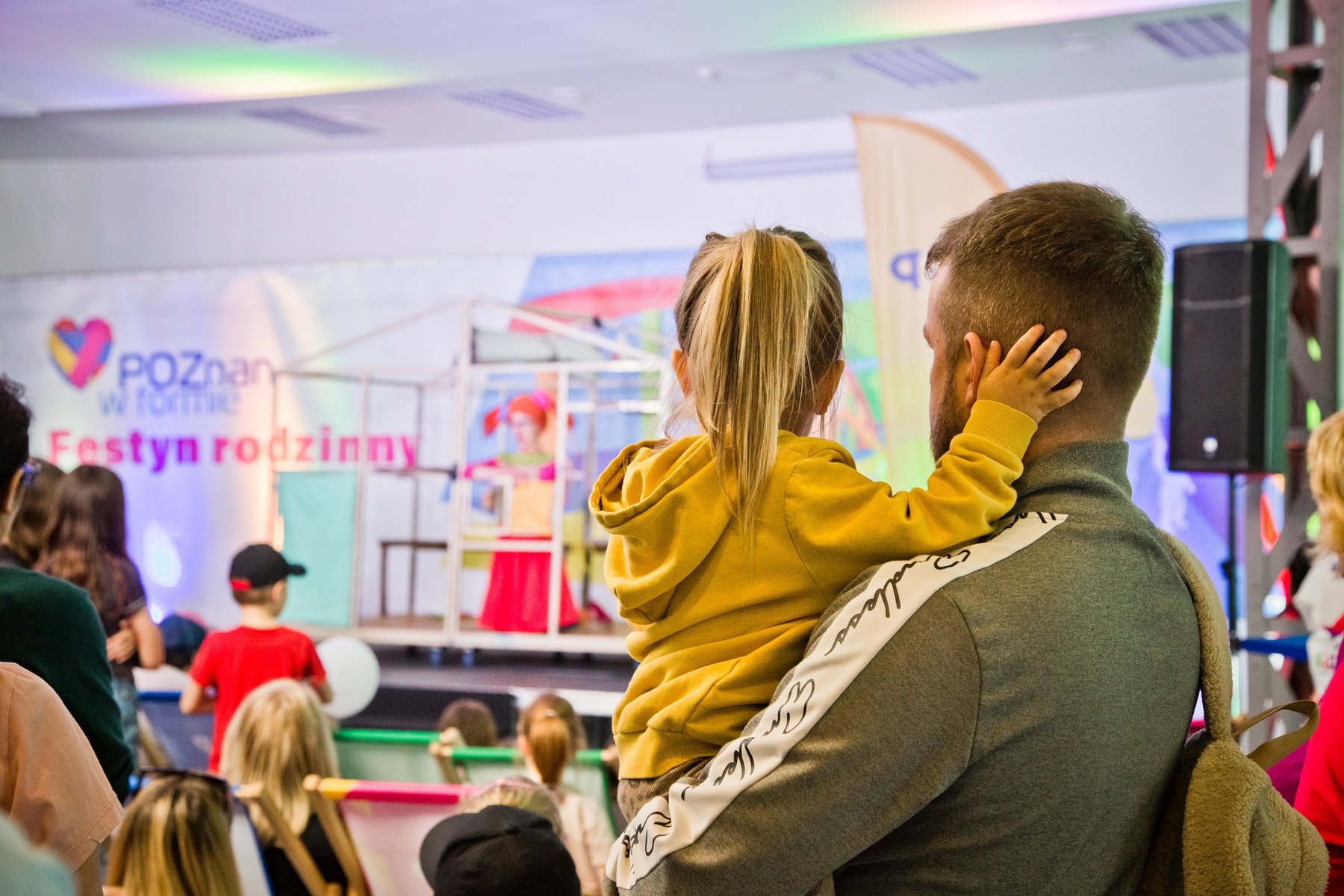 Na zdjęciu męzczyzna z dzieckiem na rękach, tyłem do obiektywu, obserwuje scenę