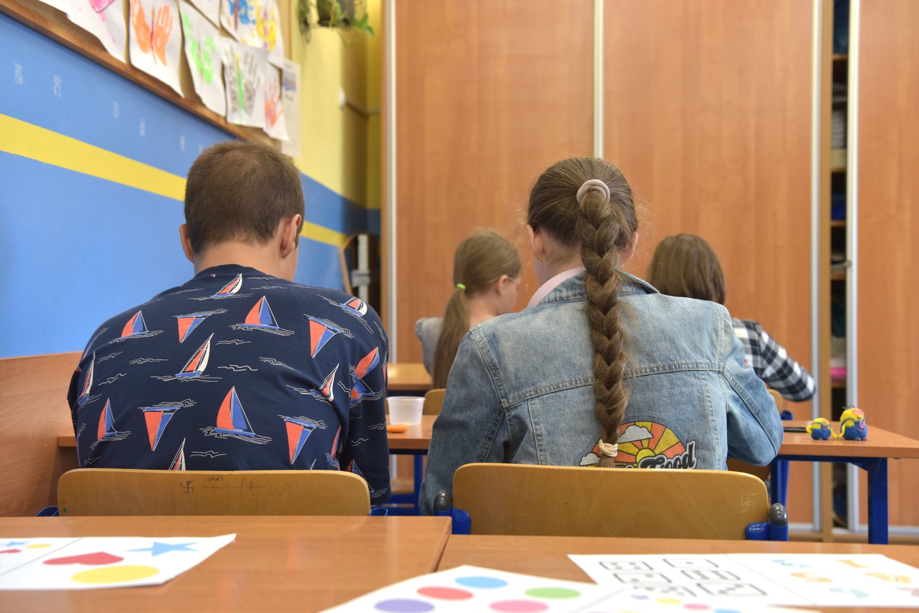 Na zdjęciu młodzi ludzie siedzący w szkolnej ławce, tyłem do obiektywu