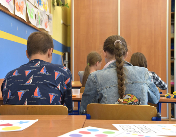 Na zdjęciu młodzi ludzie siedzący w szkolnej ławce, tyłem do obiektywu