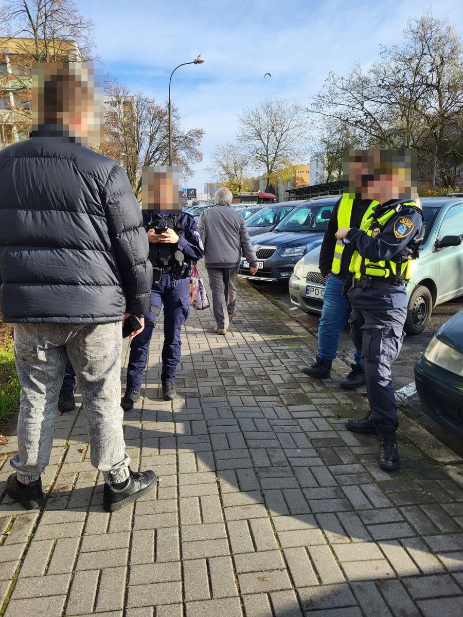 na zdjęciu na chodniku tyłem stoi mężczyzna w czarnej, pikowanej kurtce, naprzeciw niego stoi policjantka sprawdzająca w telefonie bezę danych, obok stoi strażniczka miejska i aplikant w odblaskowej kamizelce. Twarze są zanimizowane.