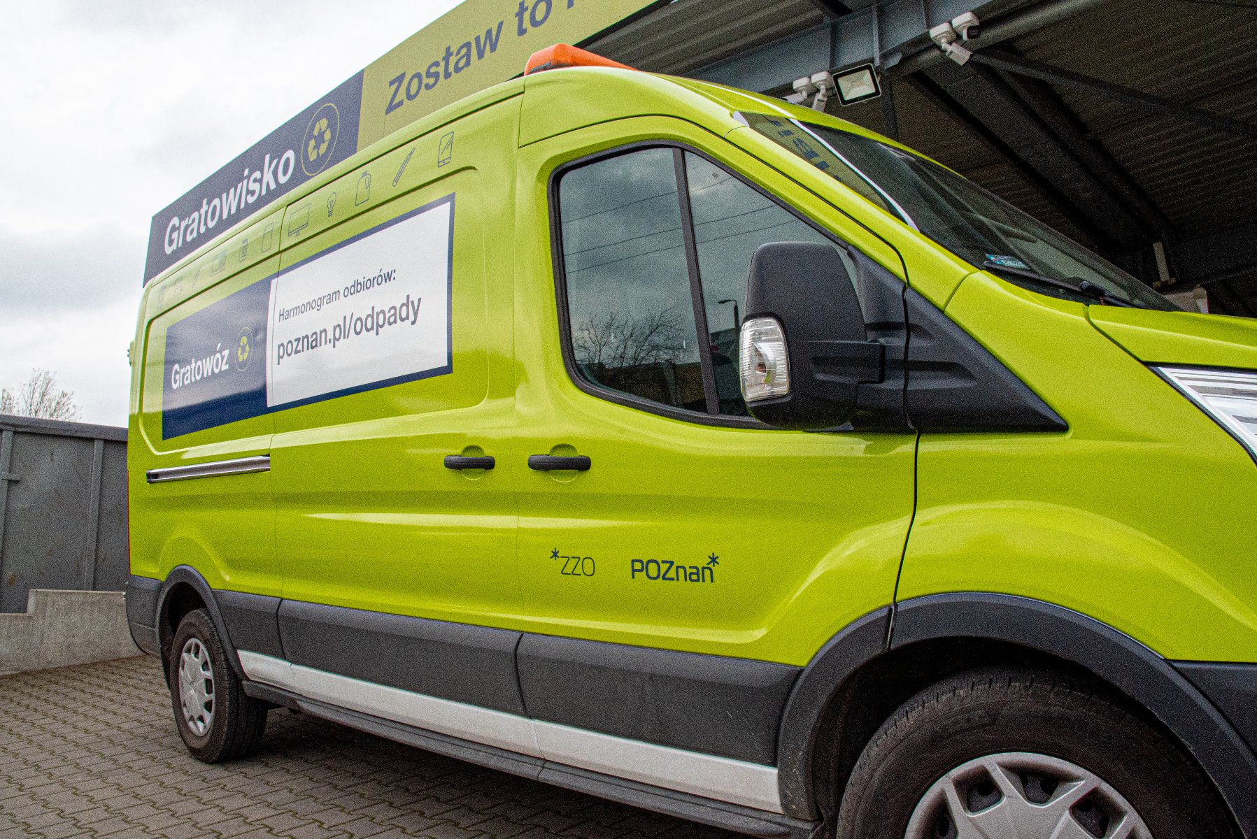 Na zdjęciu, na pierwszym planie, widać samochód typu bus, oznakowany grafikami informującymi, że odbiera on odpady od mieszkańców