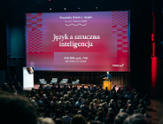 Na zdjęciu sala wypełniona publicznością, przy mównicy mężczyzna, za nim duzy ekran