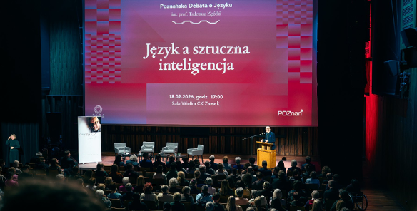 Na zdjęciu sala wypełniona publicznością, przy mównicy mężczyzna, za nim duzy ekran