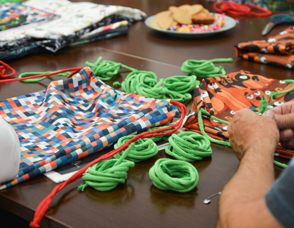 Na zdjeciu seniorki robiące plecaczki z tkaniny, widać tylko ich dłonie, w centrum zdjęcia kolorowe tkaniny i sznurki