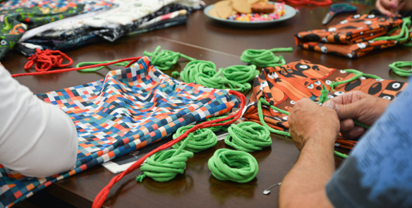 Na zdjeciu seniorki robiące plecaczki z tkaniny, widać tylko ich dłonie, w centrum zdjęcia kolorowe tkaniny i sznurki