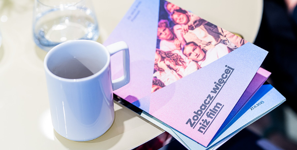 Na zdjęciu stół, na nim kubek i program festiwalu z napisem: zobacz więcej niż film