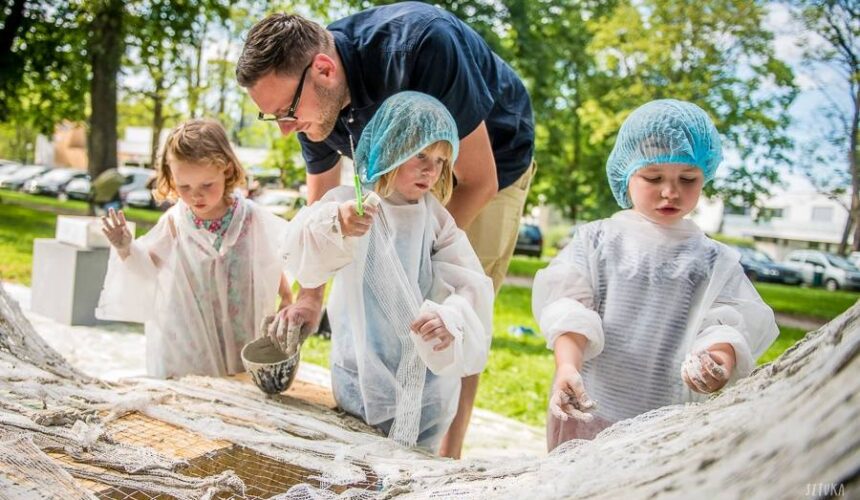 Na zdjęciu troje małych dzieci w fartuchach, nad nimi pochylony dorosły