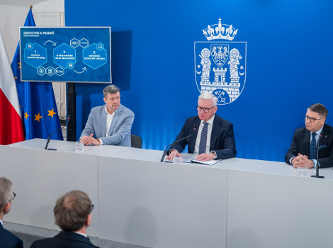 Na zdjęciu trzej mężczyźni w garniturach, siedzący za stołem podczas konferencji prasowej