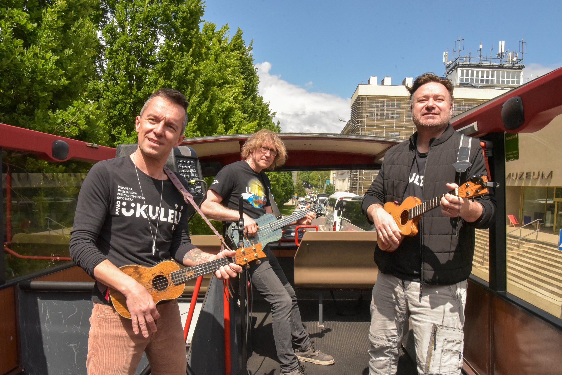 Na zdjęciu trzej mężczyźni w otwartym autobusie, grający na ukulele