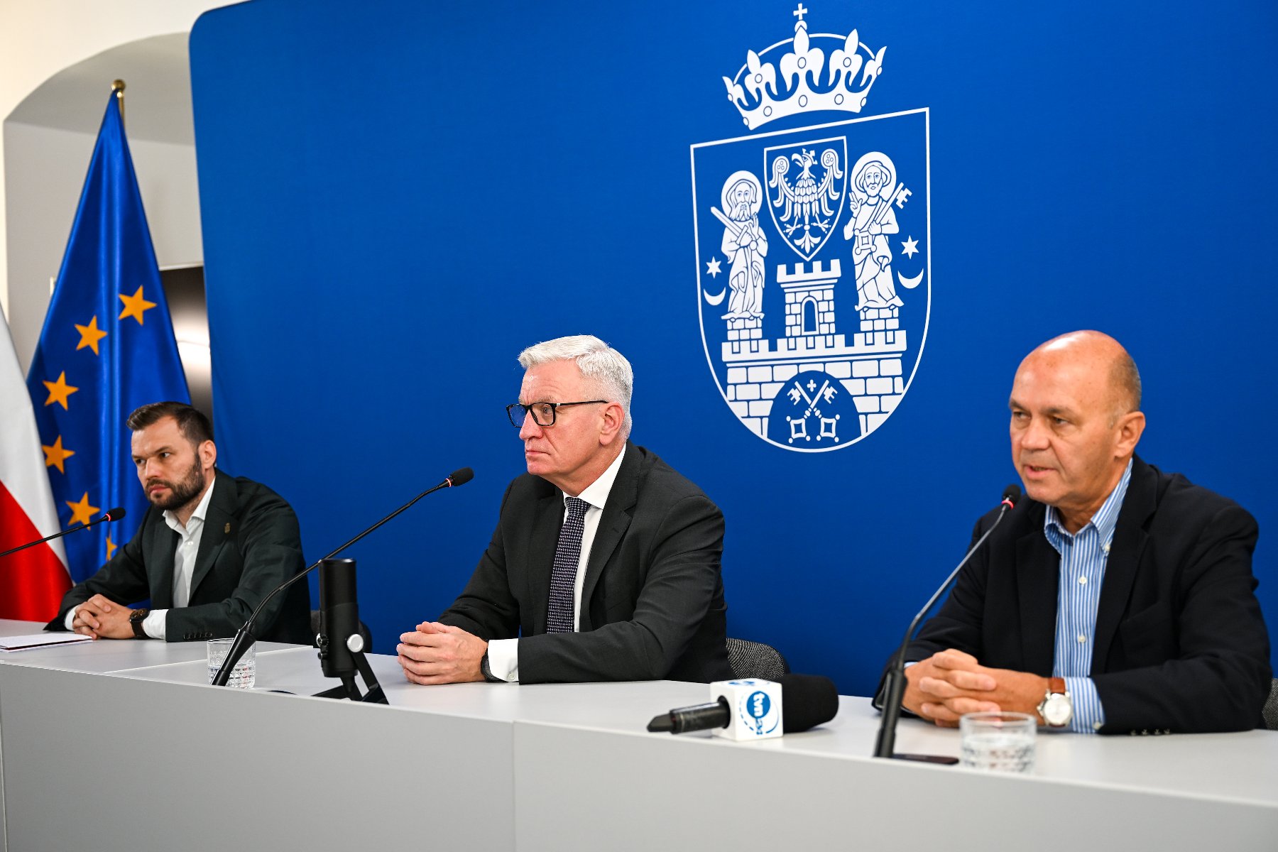 Na zdjęciu trzej mężczyźni za stołem konferencyjnym, w tle ścianka i flagi Polski oraz UE