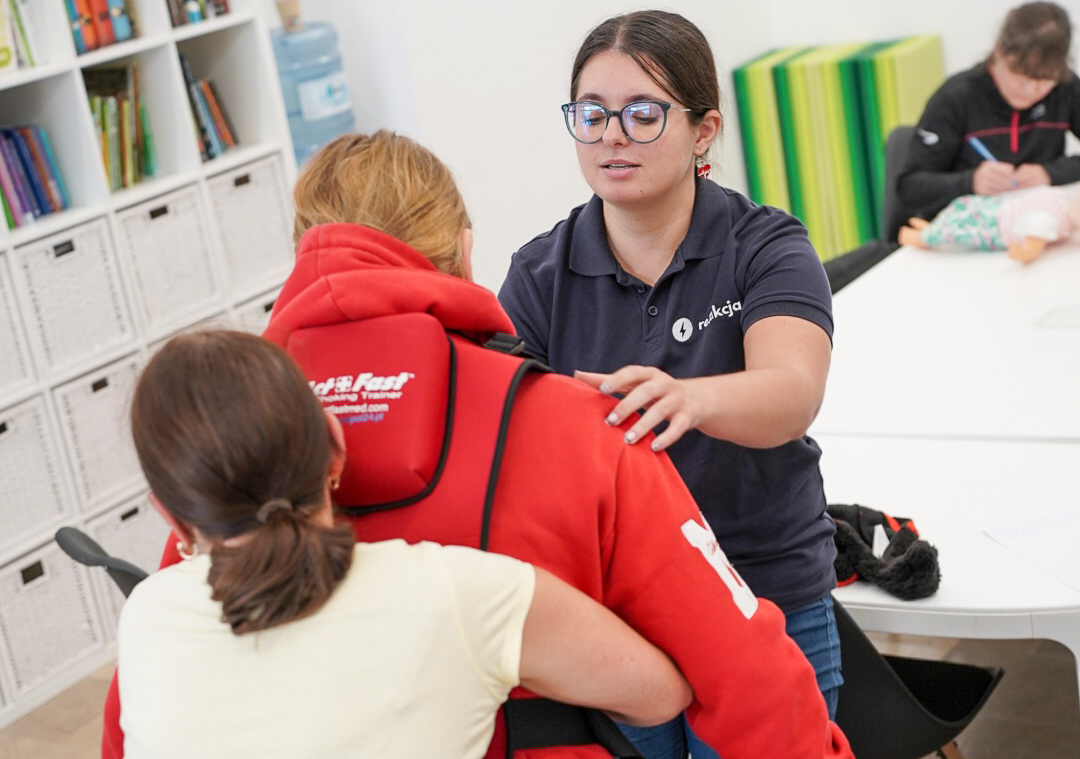 Na zdjeciu trzy osoby, jedna z nich trzyma drugą oburącz, jakby pomagała jej przy zadławieniu