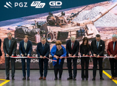 Na zdjęciu widać 9 osób stojących w rzędzie obok siebie. Wśród nich znajduje się Prezydent Miasta Poznania Jacek Jaśkowiak. Osoby wspólnie trzymają biało czerwoną wstęgę. Każdy trzyma nożyczki i wspólnie ją przecinają. W tle znajduje się duża grafika, na której widać czołg oraz dwóch żołnierzy.