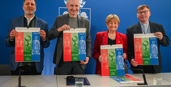 Na zdjęciu widać cztery osoby stojące za jasnym stołem na tle niebieskiej ściany z białym godłem Poznania. Każda z osób trzyma przed sobą pionowy kalendarz zatytułowany "GOŚCINNOŚĆ. Kalendarz Trzech Religii".