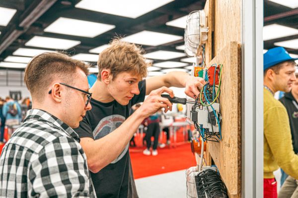 Na zdjęciu widać dwóch młodych mężczyzn pracujących przy panelu z instalacją elektryczną. Jeden z nich używa śrubokręta, koncentrując się na podłączeniu przewodów, podczas gdy drugi uważnie obserwuje. W tle znajdują się inne osoby, co sugeruje, że scena ma miejsce na targach.