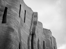 Na zdjęciu widać fragment fasady centrum handlu i sztuki Stary Browar wykonanej z pofalowanego metalu. Zdjęcie jest w kolorach czerni, bieli i szarości. W tle widać fragment nieba.