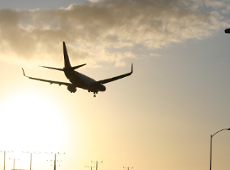 Na zdjęciu widać samolot na tle nieba. Niebo jest lekko zachmurzone. Na dole zdjęcia widać czubki słupów wysokiego napięcia oraz latarnie.