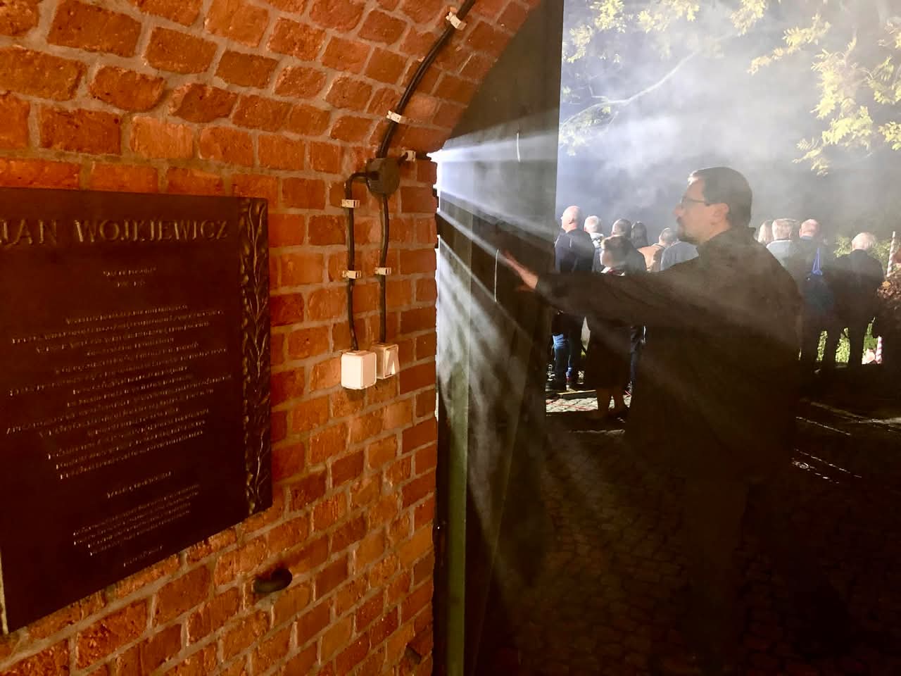 Na zdjęciu widzimy mężczyznę otwierającego metalowe drzwi osadzone w ceglanej ścianie fortu, przez które wpadają snopy światła. Po drugiej stronie drzwi, na zewnątrz, zgromadziła się grupa ludzi, którzy uczestniczą w wydarzeniu Dni Twierdzy Poznań.