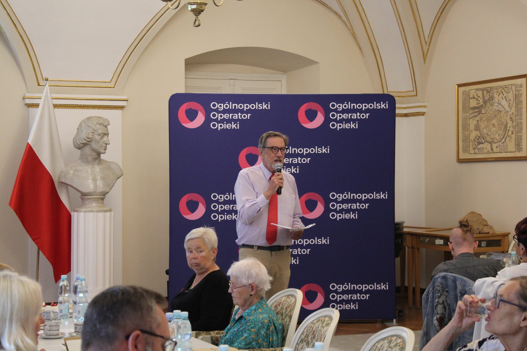 Na zdjęciu widzimy przewodniczącego MRS Pana Włodzimierza Kałka, sala Biała UMP seniorzy przy stołach, przewodniczący stoi na środku sali, w tle ścianka promocyjna Fundacji Ogólnopolski Operator Opieki.