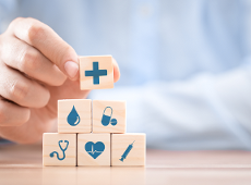 Na zdjęciu znajduje się ręka ustawiająca drewniane klocki, na których umieszczone są elementy kojarzące się ze zdrowiem, tj. stetoskop, krzyż, kropla