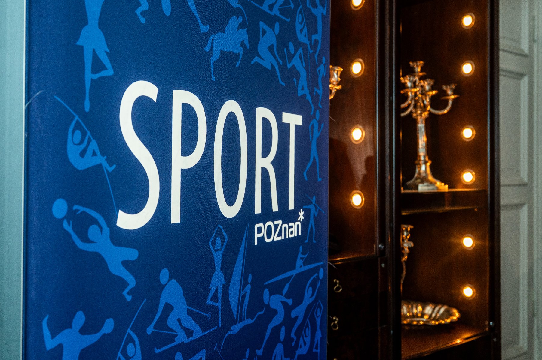 Niebieski baner z napisem "SPORT" dużymi białymi literami, poniżej napis "Poznań" z symbolem gwiazdki. Na banerze sylwetki sportowców podczas biegu, pływania i innych aktywności. Po prawej stronie oszklona witryna z ciemnego drewna, oświetlona, z prezentacją srebrnego kandelabru i tacy
